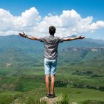 A man stands triumphantly with arms open, overlooking a scenic mountain range.